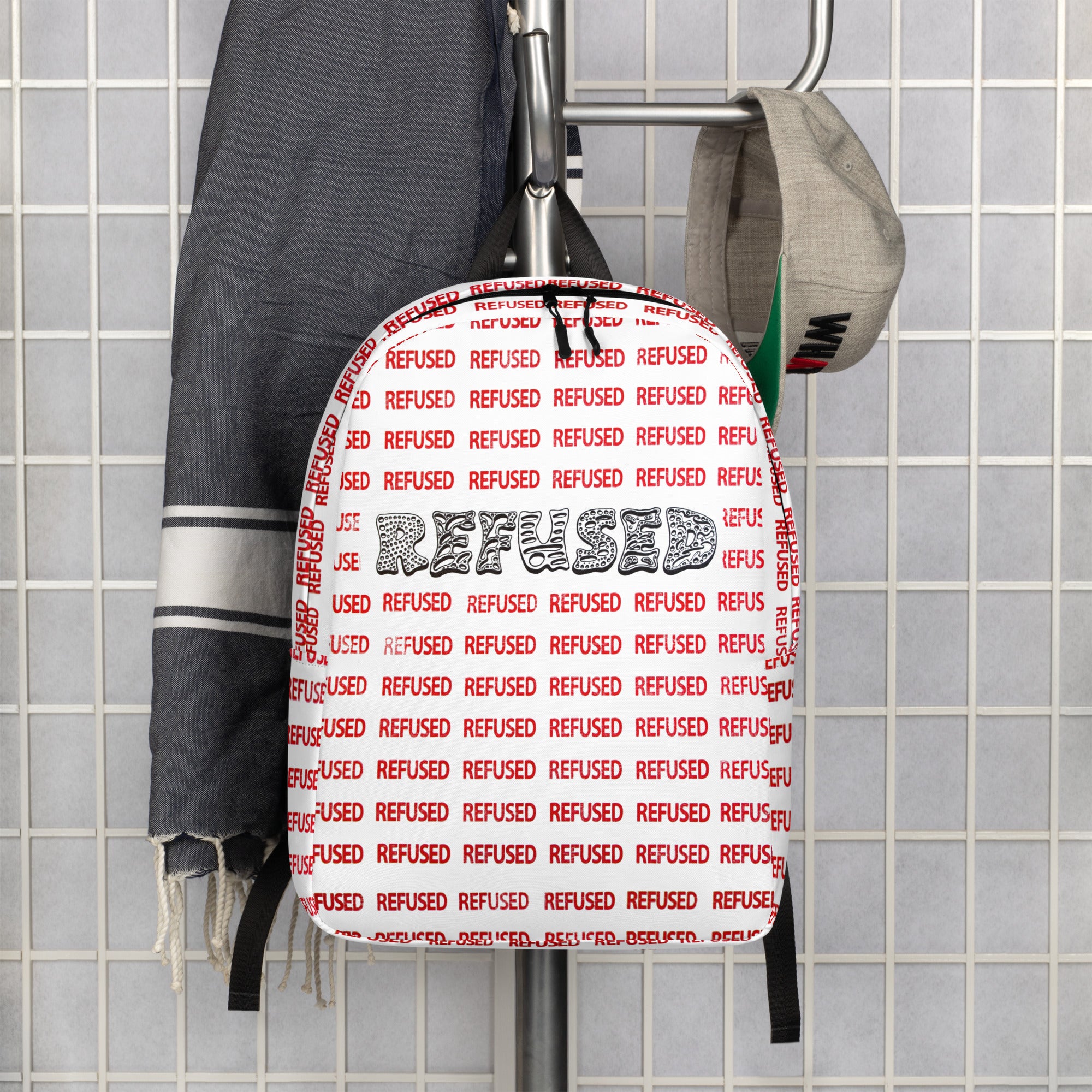 Lifestyle hanger shot of the Refused backpack—white shell with all-over red “REFUSED” print and bold black wordmark displayed against tiled wall to show modern aesthetic and scale. design by optical artist Nicholas Hayes, Chicago Illinois. 