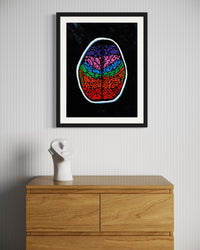 A framed artwork depicting a multicolored illustration of a human brain, displayed on a wall above a wooden cabinet. Front view of ‘Penumbra Painting No. 2’ by Nicholas Hayes. Original 18x24 inch acrylic on heavyweight canvas paper in a sleek black frame, contemporary abstract wall art, one-of-a-kind piece ready to hang.